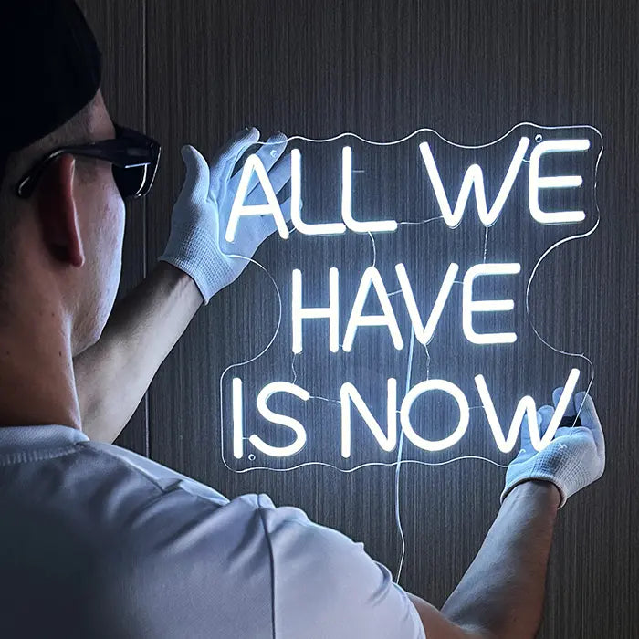 All We Have Is Now Neon LED Schild wird mit weißen Handschuhen an eine dunkle Holzwand gehalten