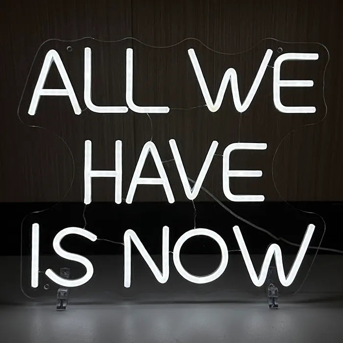 All We Have Is Now Neon LED Schild weiß leuchtend auf transparenten Standfüßen vor dunkler Holzwand