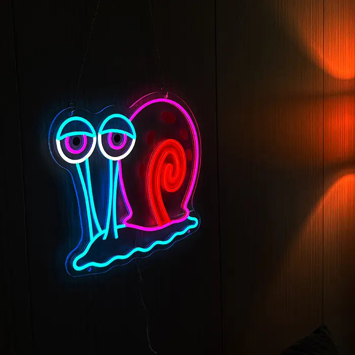 Gary the Snail Neon LED Sign hanging on a dark wooden wall with the warm light of a lamp beside it