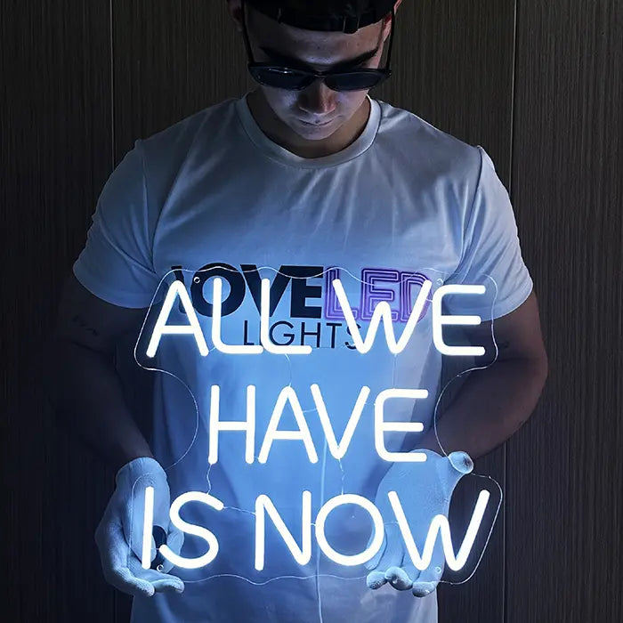 All We Have Is Now Neon LED Schild wird mit weißen Handschuhen vor dem Körper gehalten und leuchtet hell