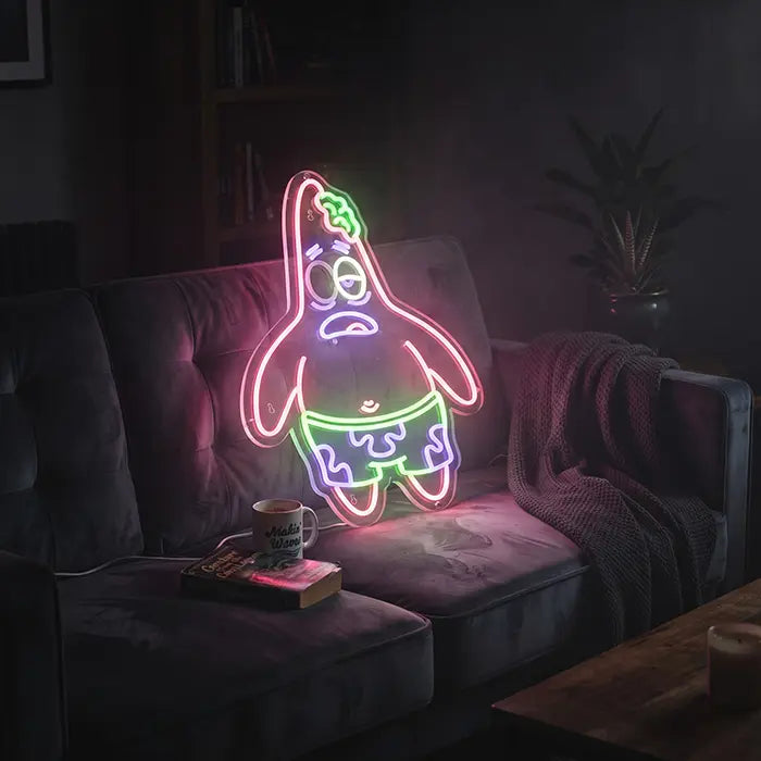 Patrick Zombie Neon LED Schild auf einem dunklen Sofa neben Tasse und Buch im abgedunkelten Wohnzimmer