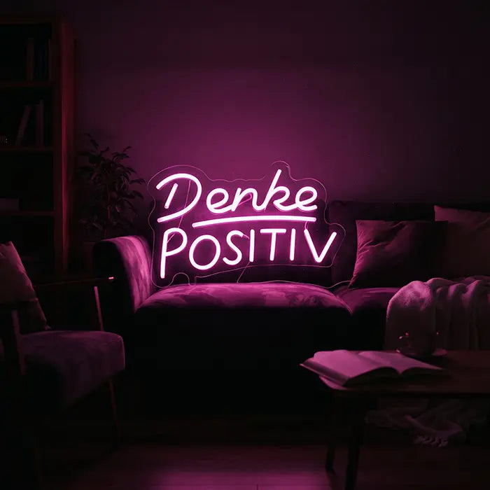 Denke Positiv Neon LED Schild rosa leuchtend an der Wand über einem Sofa im abgedunkelten Wohnzimmer