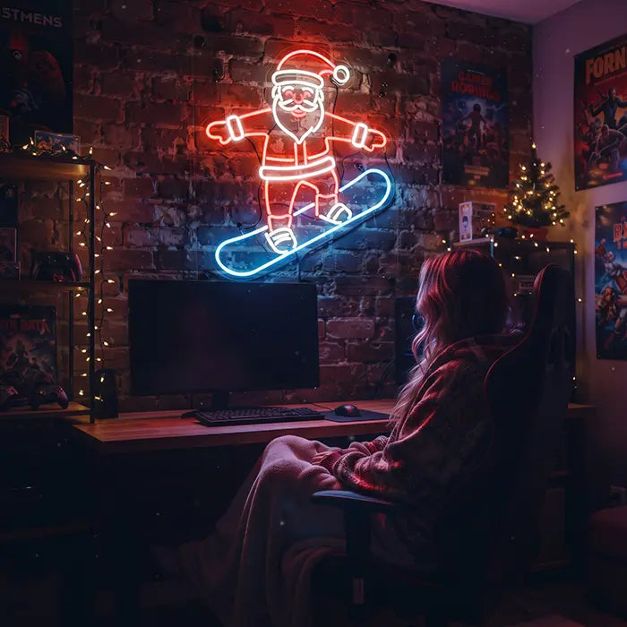 Snowboarding Santa Neon LED Sign above a gaming desk, a person in the chair in front, a Christmas tree beside