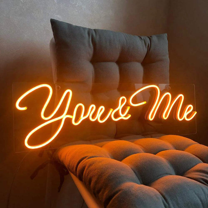 Neon LED-skilt You & Me i oransje lysende skriveskrift på en klar akrylplate foran en vegg